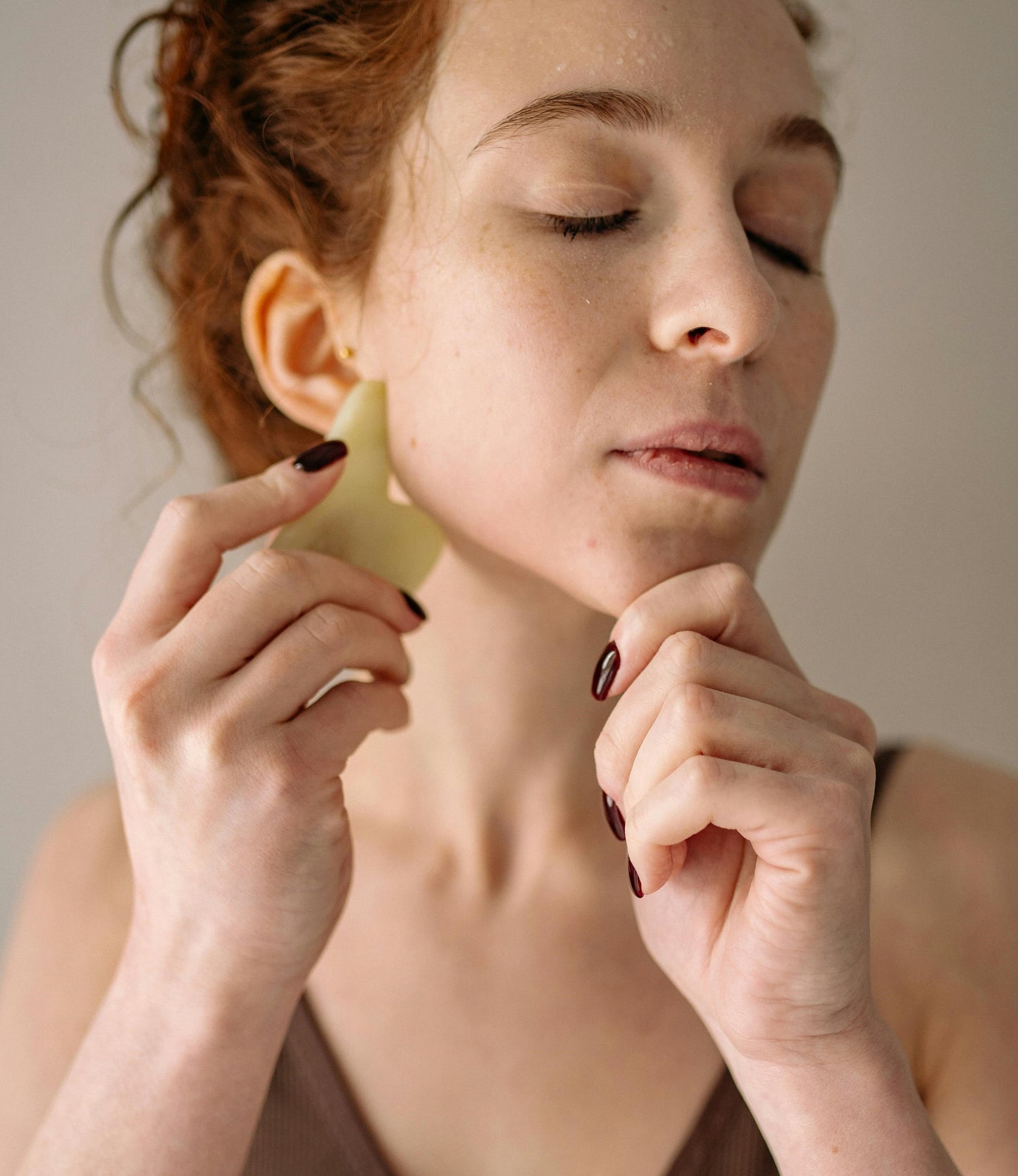 A woman with fair skin and red hair using a gua sha facial massage tool along her jawline, with a relaxed expression and closed eyes.